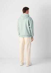Uomo di spalle che indossa un hoodie verde chiaro, pantaloni cargo color crema e sneakers bianche su uno sfondo bianco semplice.