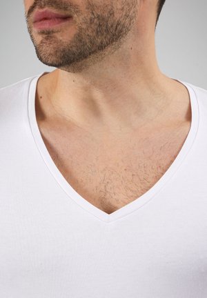 Close-up van het onderste deel van het gezicht en de nek van een man die een wit V-hals T-shirt draagt, met een korte baard en zichtbare borsthaar boven het T-shirt.