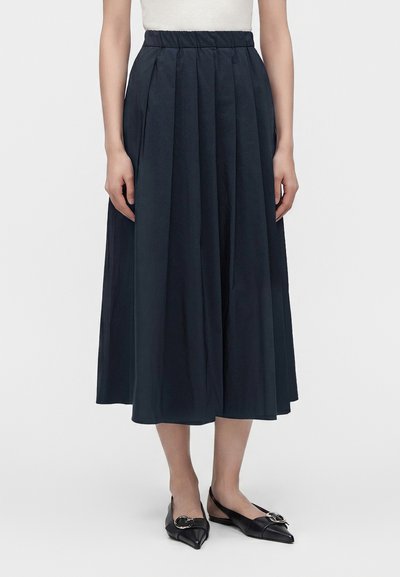 Femme portant une jupe midi plissée noire taille haute associée à des ballerines slingback noires pointues avec détail boucle en métal.