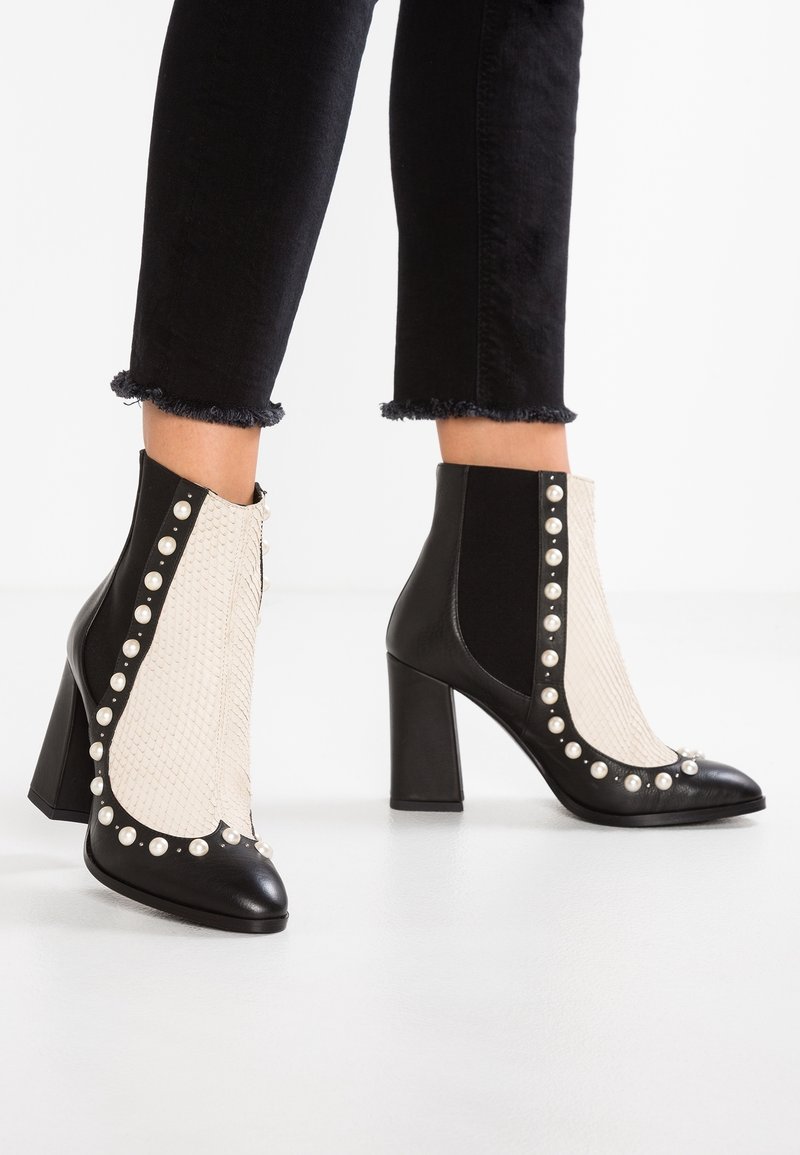 Bottes noires et blanches à talons hauts jusqu'à la cheville avec des détails en perles, portées avec un pantalon noir à ourlet effiloché sur fond blanc.