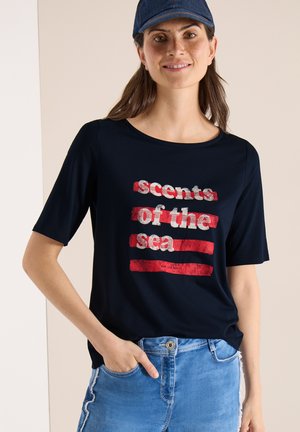 Femme portant un t-shirt bleu marine avec des rayures rouges et un texte blanc "scents of the sea", assorti à un jean bleu clair et une casquette bleu foncé.