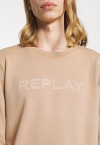 Felpa marrone chiaro con la scritta "REPLAY" in trasparenza sul petto, indossata da una persona con capelli biondi lunghi fino alle spalle.