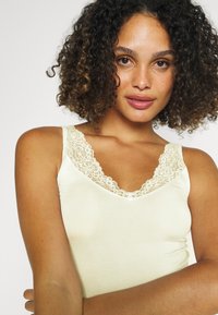 Camicetta leggera gialla chiara con un bordo in pizzo lungo il colletto e le spalline, dalla texture morbida e con un piccolo fiocco decorativo al centro.