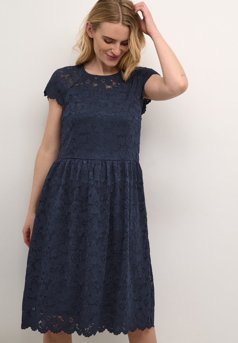 Lace dress in navy blue with a fitted bodice, short scalloped sleeves, and a flared skirt featuring a floral pattern and a scalloped hem.