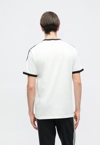 Biela tričko s čiernym rebrovaným výstrihom a manžetami na rukávoch, s krátkymi rukávmi a voľným strihom, vyrobené z hladkého bavlneného materiálu.