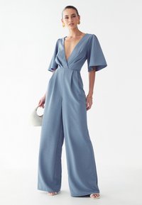 Modra kombinezonka z globokim V-izrezom, kratkimi razširjenimi rokavi, širokimi hlačami, teksturirano blago in usklajenimi dodatki, ponuja elegantno silhueto.