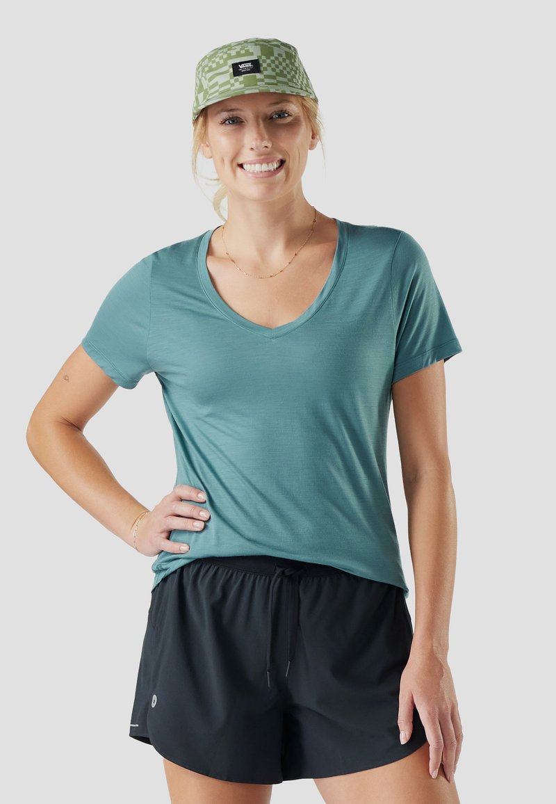 Donna sorridente che indossa un berretto a quadri verde, una maglietta a V color teal e shorts sportivi neri, con una mano sul fianco, davanti a uno sfondo grigio.