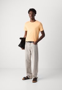 Tricou din bumbac de culoare portocalie deschisă, blugi din denim gri deschis, curea neagră și sandale negre. Modelul ține o jachetă neagră pe braț.