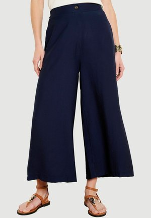 Pantaloni blu navy a gamba larga e taglio corto con vita alta, bottone nero, indossati con sandali piatti color sabbia con dettagli decorativi dorati.