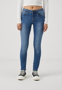 Vaqueros ajustados de color azul en un lavado medio con cintura de altura media, que cuentan con bolsillos frontales y una textura suave. Llevados con zapatillas negras y blancas.