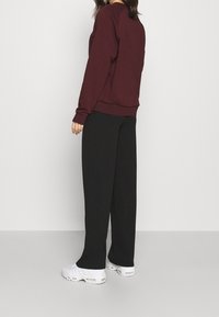 Sweat-shirt bordeaux avec col rond et poignets côtelés, associé à un pantalon large noir et des baskets blanches, photographié de dos.