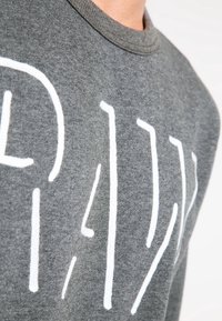 Sudadera gris hecha de una tela suave, con un diseño de texto blanco "RAW" prominente en una fuente estilizada en el frente. Cuello redondo.