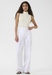 Bijele, široke hlače s plitkim naborima uparene s uskim, bezrukavim, krem turtleneck puloverom. Glatka, lagana tkanina; minimalistički dizajn.