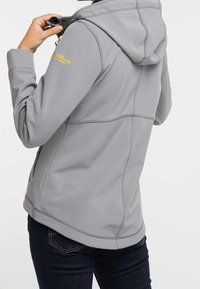 Osoba upravující kapuci na světle šedé bundě s žlutým vyšitým textem na rukávu, oblečená v tmavě modrých džínách.