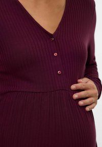 Robe longue à manches longues bordeaux côtelée avec un décolleté en V et quatre détails en bouton sur le devant. Taille élastique avec des plis subtils en dessous.