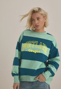 Zeleno-modrá pruhovaná mikina s nápisem "PORTLAND Oregon" v žlutém písmu, s kulatým výstřihem a volnými rukávy.