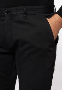 Zwarte broek van gladde stof met een getailleerde pasvorm, voorzien van een knoopsluiting en voorkanten zakken aan elke kant.