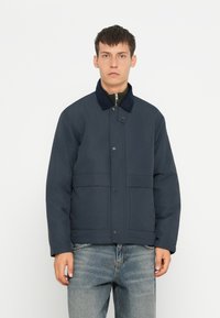 Giacca imbottita di colore blu navy con chiusura a zip, bottoni a pressione, due tasche anteriori e colletto scuro a contrasto. Texture liscia.