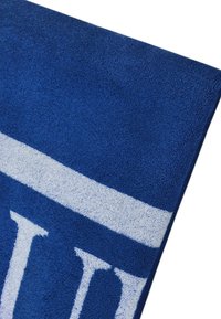 Asciugamano in spugna blu con una finitura morbida e testurizzata. Presenta ampie strisce bianche e lettere; la vista angolata rivela i dettagli del tessuto e il contrasto dei colori.