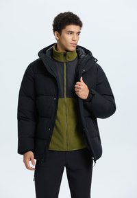 Zwarte pufferjack met capuchon, voorzien van een contrasterende groene fleece met rits eronder. De jas heeft een gegokt ontwerp en een ritssluiting aan de voorkant.