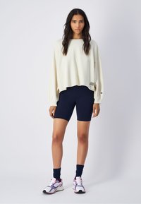 Sweatshirt blanc court à manches longues, associé à un short cycliste bleu marine. Chaussures de sport blanches et violettes avec des chaussettes bleu foncé.