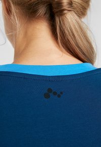 Dunkelblaues T-Shirt mit einem hellblauen Kragen, das auf der Rückseite ein schwarzes kreisförmiges Motiv aufweist, aus einem weichen, glatten Stoff gefertigt.