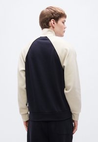 Ανδρικό φούτερ με ψηλό γιακά, μπλοκ χρωμάτων σε μαύρο και κρεμ, απαλό ύφασμα και μανίκια ράγκλαν για άνετη εφαρμογή.