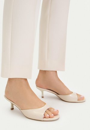 Pies con mulettes de tacón bajo color crema y punta abierta, combinadas con pantalones cortos beige claro sobre un fondo liso.