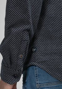 Marineblå mønstret skjorte med en struktureret vævning, der har en knapmanchet og et lille mærke, båret sammen med lyse denimjeans.