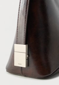 Κοντινό πλάνο γωνίας τσάντας από σκούρο καφέ δέρμα με ραμμένο λουράκι και ασημί ορθογώνιο μεταλλικό κούμπωμα που δείχνει το όνομα της μάρκας.