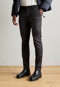 Homme portant un pantalon slim noir, des bottines Chelsea en cuir noir, une chemise blanche et une veste matelassée navy, se tenant sur un sol en bois.