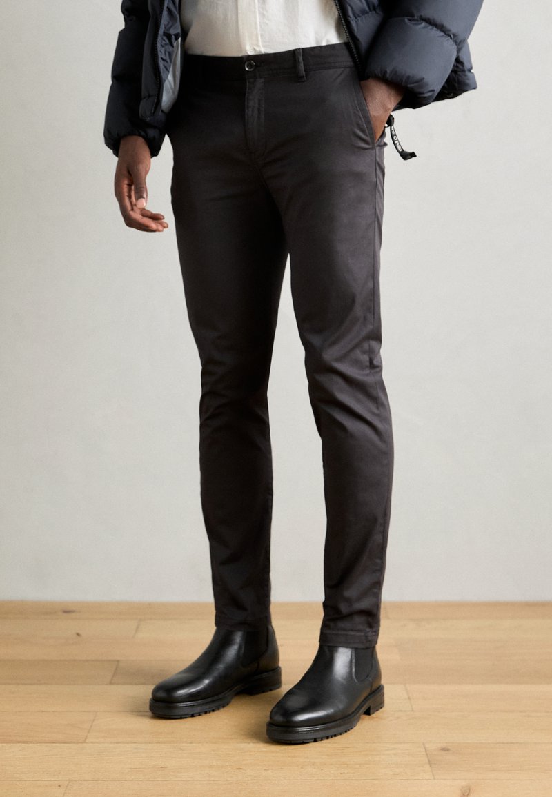 Homme portant un pantalon slim noir, des bottines Chelsea en cuir noir, une chemise blanche et une veste matelassée navy, se tenant sur un sol en bois.