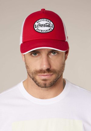 Man met baard, draagt een rood-witte Coca-Cola honkbalpet en een wit shirt, neutrale achtergrond, kijkt recht vooruit.