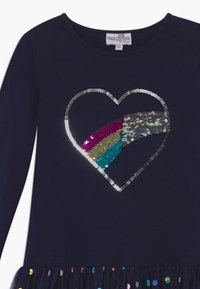 Námořnické šaty s dlouhým rukávem, které mají stříbrné flitrové srdce s barevnými proužky a volánkový lem ozdobený mnohobarevnými tečkami.