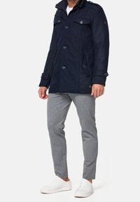 Man wearing navy blue buttoned coat, gray slim-fit pants, and white sneakers, standing against a plain light background.