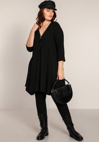 Robe tunique noire à côtes avec un décolleté en V et une fermeture éclair, associée à des leggings noirs et des bottes de combat. Les accessoires comprennent un sac à main texturé noir.