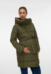 Kvinde i olivengrøn belagt dunjakke med høj krave, som holder sig om maven, iført blå jeans og dråbeøreringe foran en hvid baggrund.