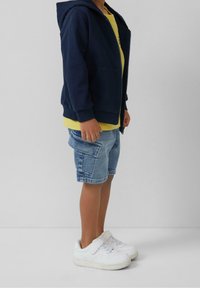 Námořnická šedá mikina na zip, žluté tričko, džínové cargo šortky, bílé tenisky. Důraz na pohodlí a volný střih. Krátké rukávy, elastické manžety.