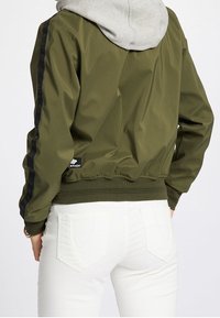 Olivgrön bomberjacka med ribbad nederkant, prydd med svarta sidostråk och en logotypapplikation. Grå huva som stickar fram under.