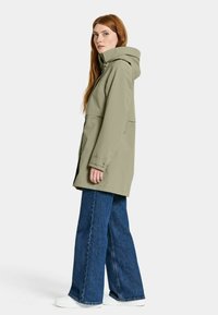 Vihreä vettähylkivä takki hupulla, jossa on vetoketjukiinnitys edessä, sivutaskut ja rento malli, yhdistettyinä leveälahkeisiin farkkuihin.