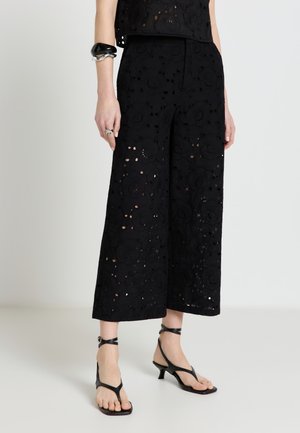 Pantaloni a gamba larga in pizzo floreale nero, abbinati a sandali neri con cinturini e tacco basso e braccialetti argento e neri al polso sinistro.