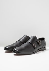 Scarpe eleganti in pelle nera con doppia fibbia a monk strap, punte affusolate e tacco basso su una superficie bianca.