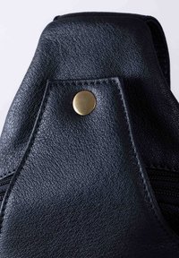 Black leather bag with a textured finish, featuring a prominent gold snap button and contrasting stitching along the edges. Zipper visible.