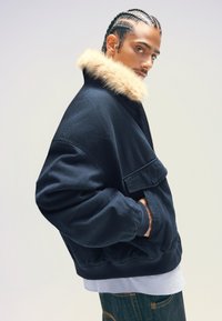 Marinblå bombarjacka med pälsfodrad krage, med dragkedja framtill och stora fickor. Tillverkad av mjukt material med en något oversized passform.