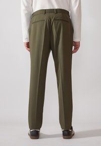 Olivgrüne maßgeschneiderte Hose mit geradem Bein, Gürtelschlaufen und zwei Gesäßtaschen mit dezenten Paspelierungen.