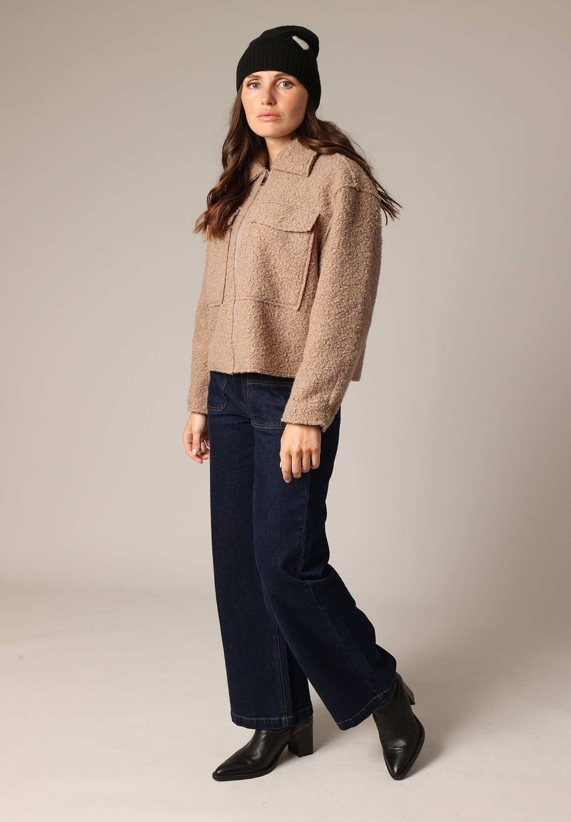 Femme portant une veste zippée beige texturée, un jean large noir, des bottes à talons noires et un bonnet en tricot noir, se tenant sur un fond neutre.