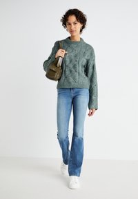 Green knitted sweater with a textured, cable design and ribbed cuffs, paired with light blue flared jeans and white sneakers, holding a small bag.