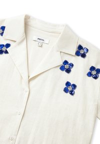 Camisa de manga corta color crema con cuello, adornada con flores de lentejuelas azules y acentos de cristales en los hombros. Tejido texturizado.