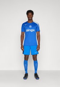 Blå Nike fodboldoutfit med en T-shirt med korte ærmer og hvide detaljer, shorts, knælange strømper og sorte støvler.