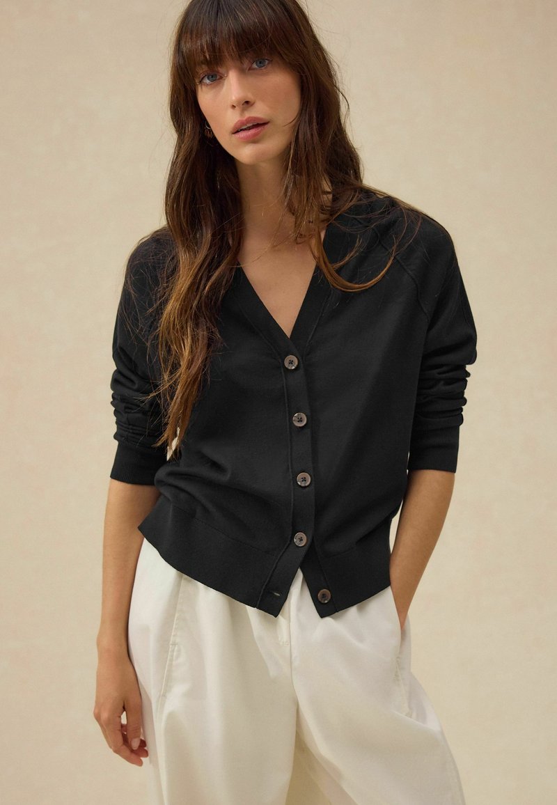 Cardigan noir avec un col en V, sept boutons marron et des poignets côtelés. Porté sur un pantalon blanc, présentant une texture lisse et une coupe ajustée.